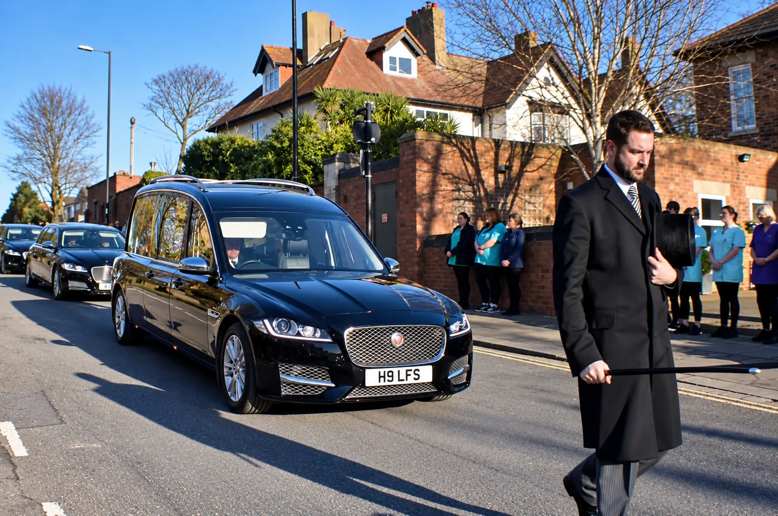 A funeral director carrying his top hat walks ahead of the Jaguar hearse